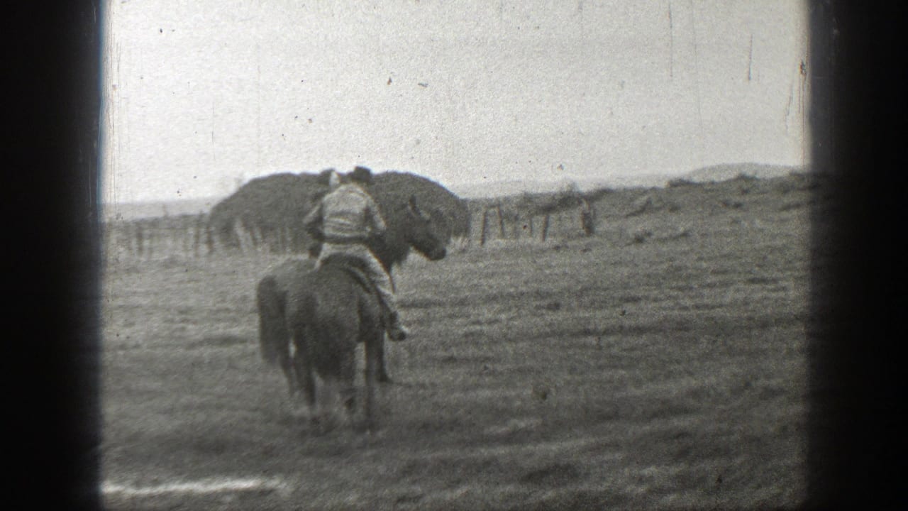Two mounted riders travel away across an open field with a fence in the distance.