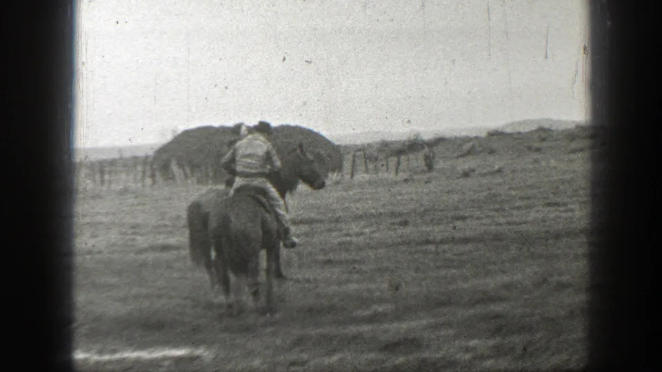 Two riders on horseback move through a muddy Montana field, with a fence line and low rural landscape in the background.