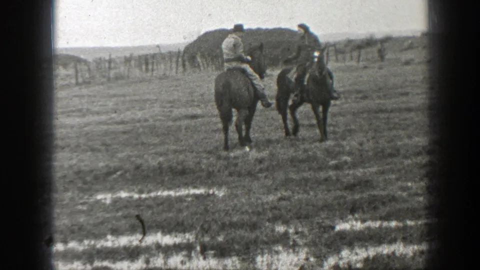 In a rural Montana field, two mounted riders appear to slow or pause near puddled ground and a fence line.