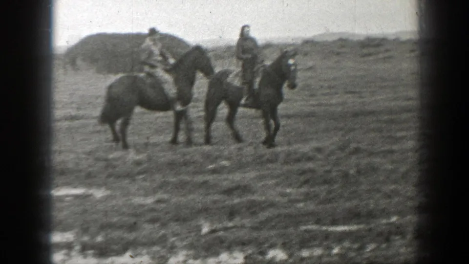 Two people on horseback face each other in the muddy field, framed by soft 8mm grain and a cloudy rural background.