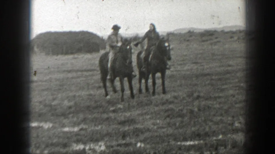 The riders approach the camera on horseback in rural Montana, with a low hill behind them.