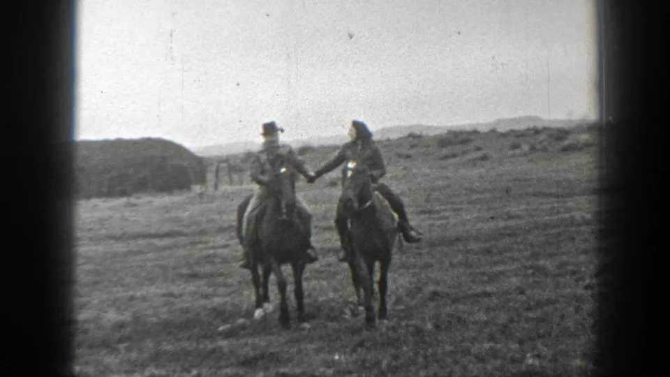 Two adults on horseback ride side by side across a rural Montana field, framed by 8mm film edges.