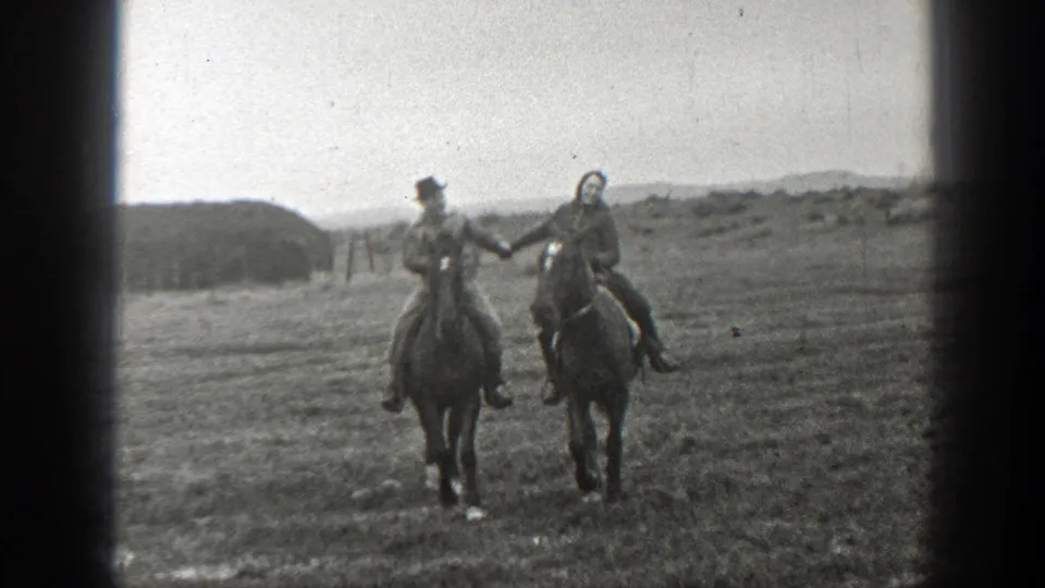 In Montana 1939 home-movie footage, two adults on horseback ride side by side across an open plain, appearing to hold hands.