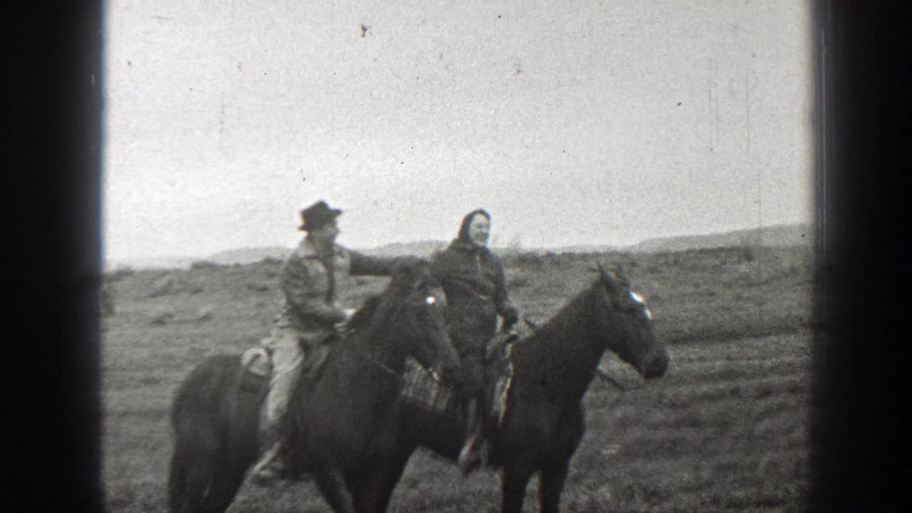 Two mounted adults ride close together, one wearing a brimmed hat, on an open plain.