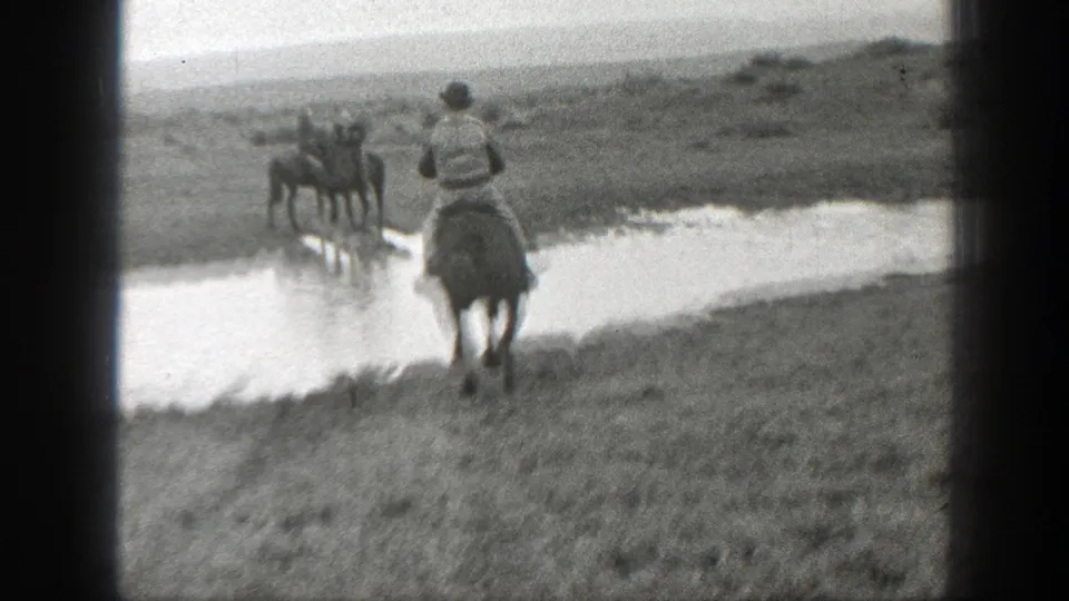 1939 Montana 8mm frame showing a man on horseback crossing open ground near a muddy waterway, with another rider farther away.