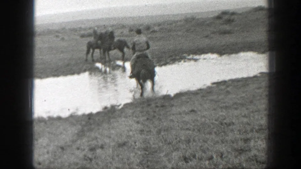 Men on horseback appear to cross a shallow stream in Montana grassland, filmed in soft 8mm black-and-white footage.