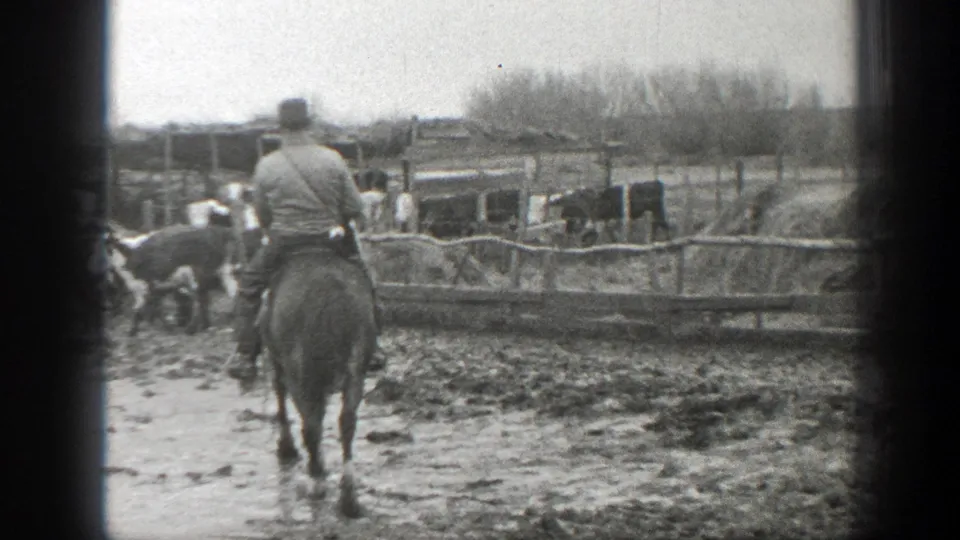 A mounted rancher in a wide-brimmed hat rides through a muddy corral area beside wooden fencing in Montana, 1939.