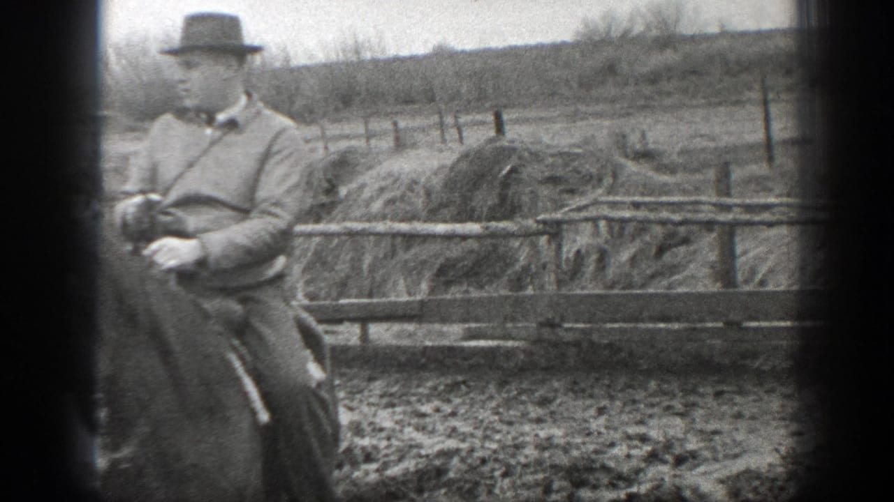 In 1939 Montana home-movie footage, a man rides a horse past fence rails and hay or straw stacks in a muddy farmyard.