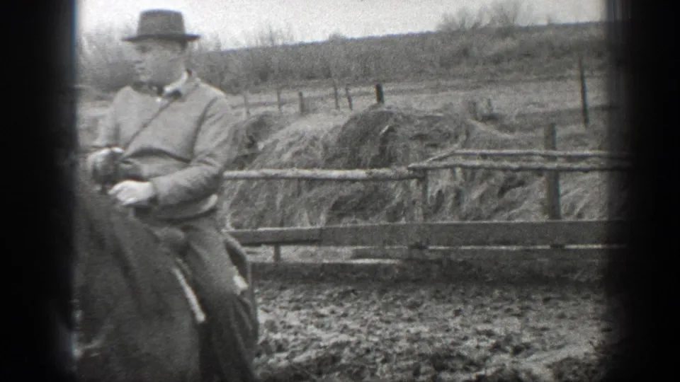 In 1939 Montana home-movie footage, a man rides a horse past fence rails and hay or straw stacks in a muddy farmyard.