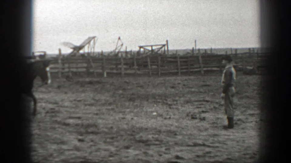 A man in period work clothes stands in a fenced Montana farm paddock while a horse moves nearby.
