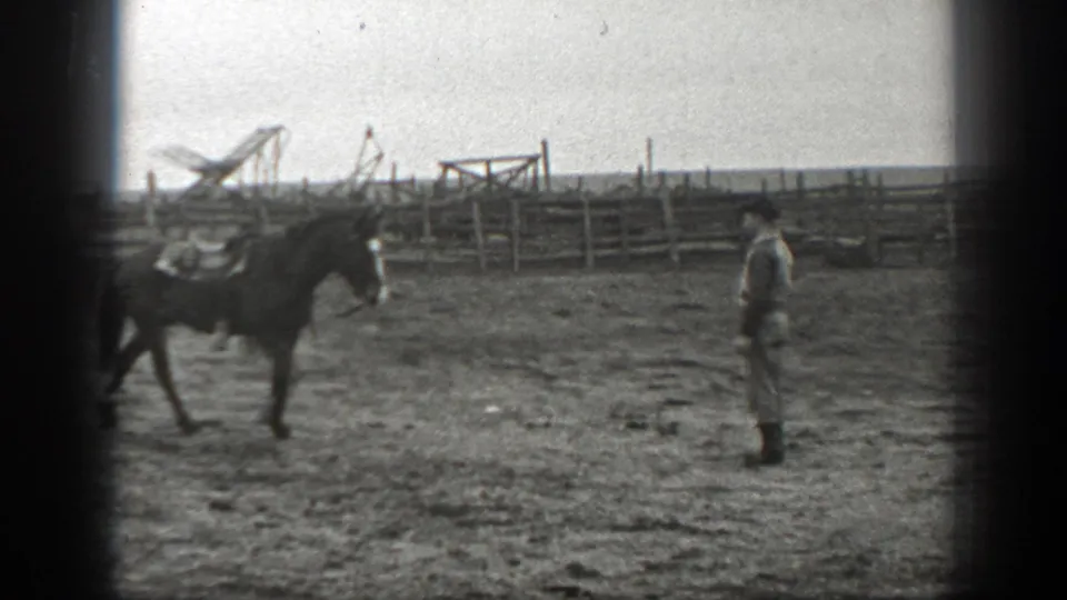 A saddled horse walks past a man in a rural Montana paddock, with fences and farm equipment in the background.