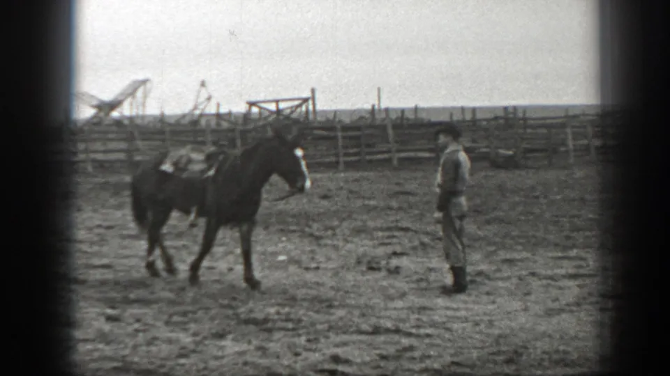 In 1939 Montana home-movie footage, a saddled horse approaches an adult man in a dark hat inside a fenced dirt corral.