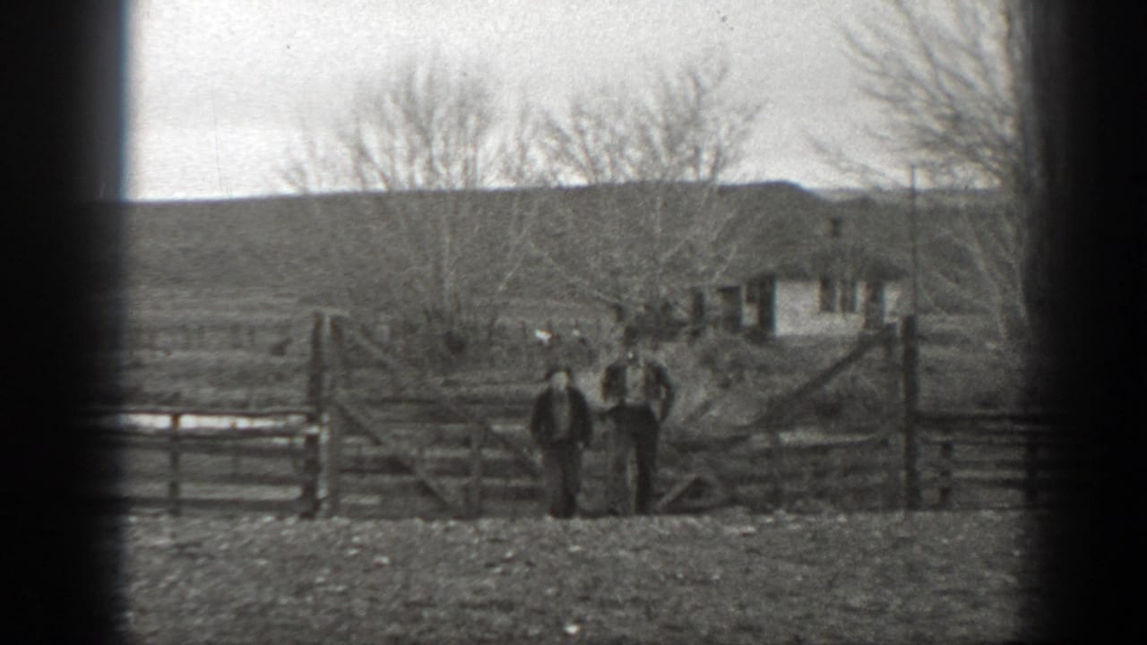 Two people in rural work clothing stand by a wooden gate on a Montana ranch, with open land, bare trees, and a small building in the background.