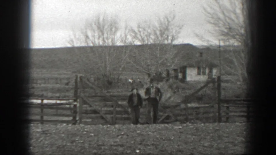Two people in rural work clothing stand by a wooden gate on a Montana ranch, with open land, bare trees, and a small building in the background.