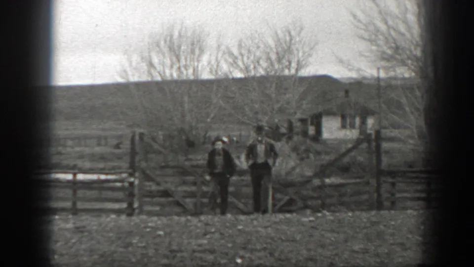 Two people remain near the wooden ranch gate, with fencing, bare trees, a small building, and open Montana countryside behind them.