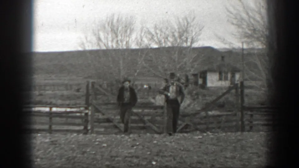 Two figures stand near a wooden ranch fence and gate in black-and-white 8mm footage from Montana, with bare trees and a small building beyond.