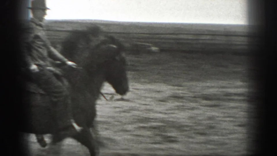 A rider in a hat passes across a dusty ranch area on horseback, with open fencing and flat Montana countryside in the background.