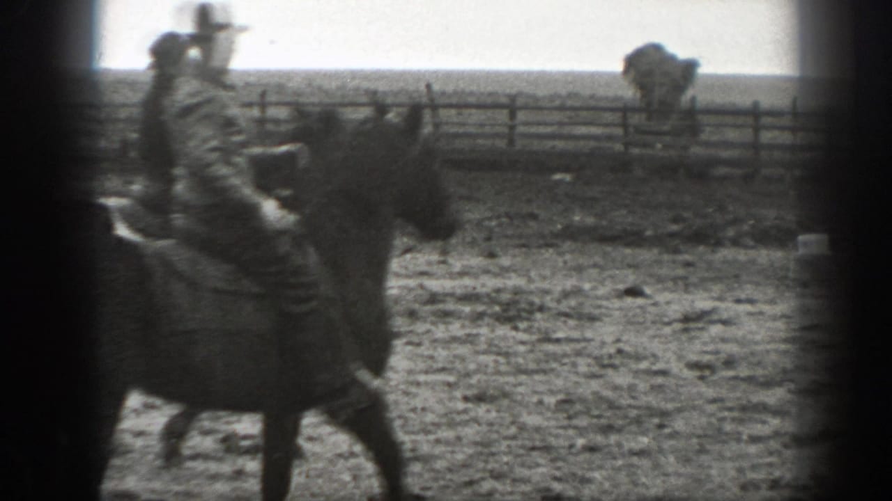 Man in a hat rides a dark horse through a fenced dusty field.