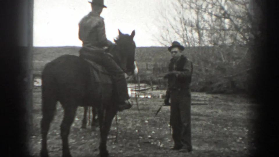 A mounted rider pauses near another adult man standing by sparse trees in rural Montana, 1939.