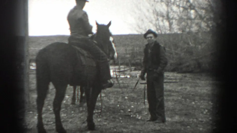 1939 Montana ranch scene with a standing man in a dark hat near a saddled horse and rider, framed by bare trees and pasture.