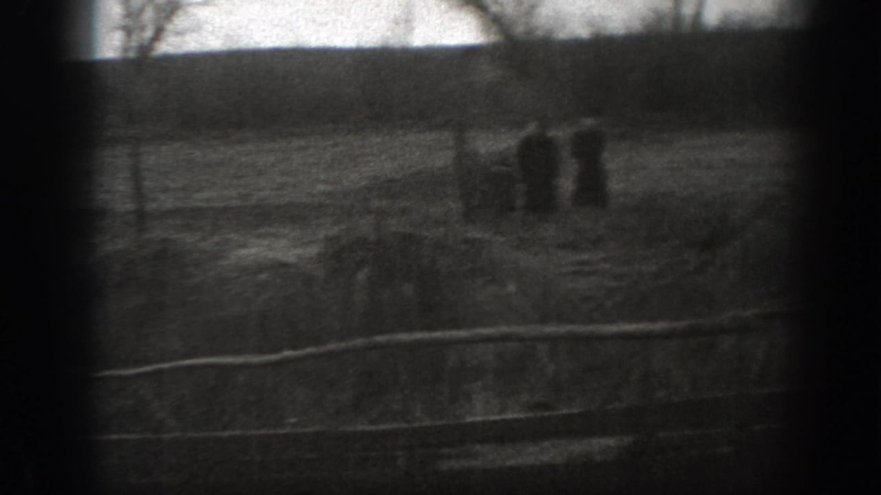 Grainy 1939 Montana 8mm frame of a fenced pasture with distant cattle forms, open grass, and a dark tree line beyond.