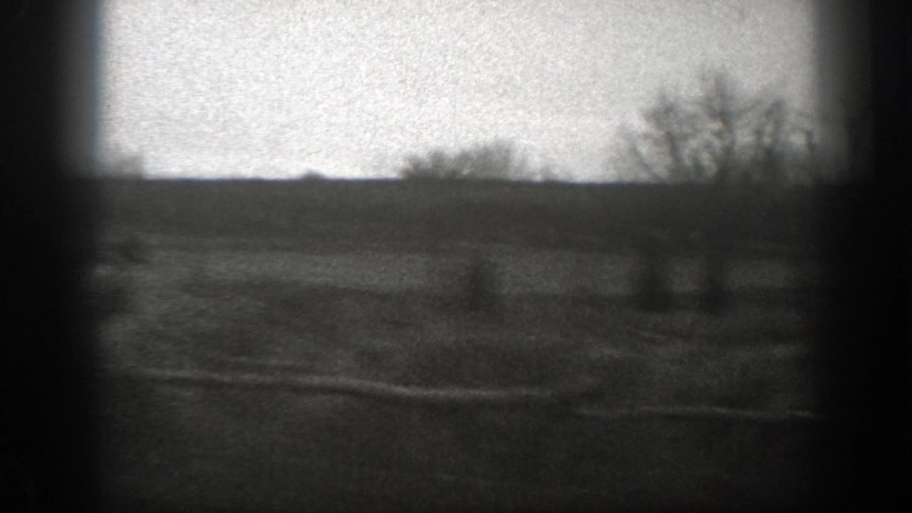 Grainy 1939 Montana 8mm frame of open farmland with rough fence rails, bare trees, and a distant low horizon.