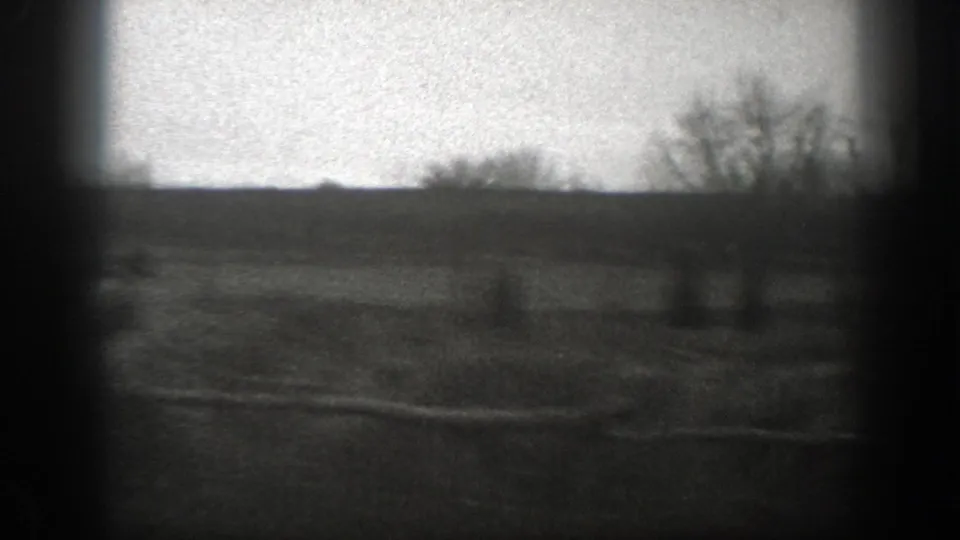 Grainy 1939 Montana 8mm frame of open farmland with rough fence rails, bare trees, and a distant low horizon.