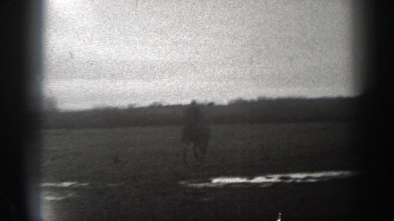 1939 Montana 8mm frame showing a soft, distant rider on horseback crossing a broad field with low hills or treeline in the background.