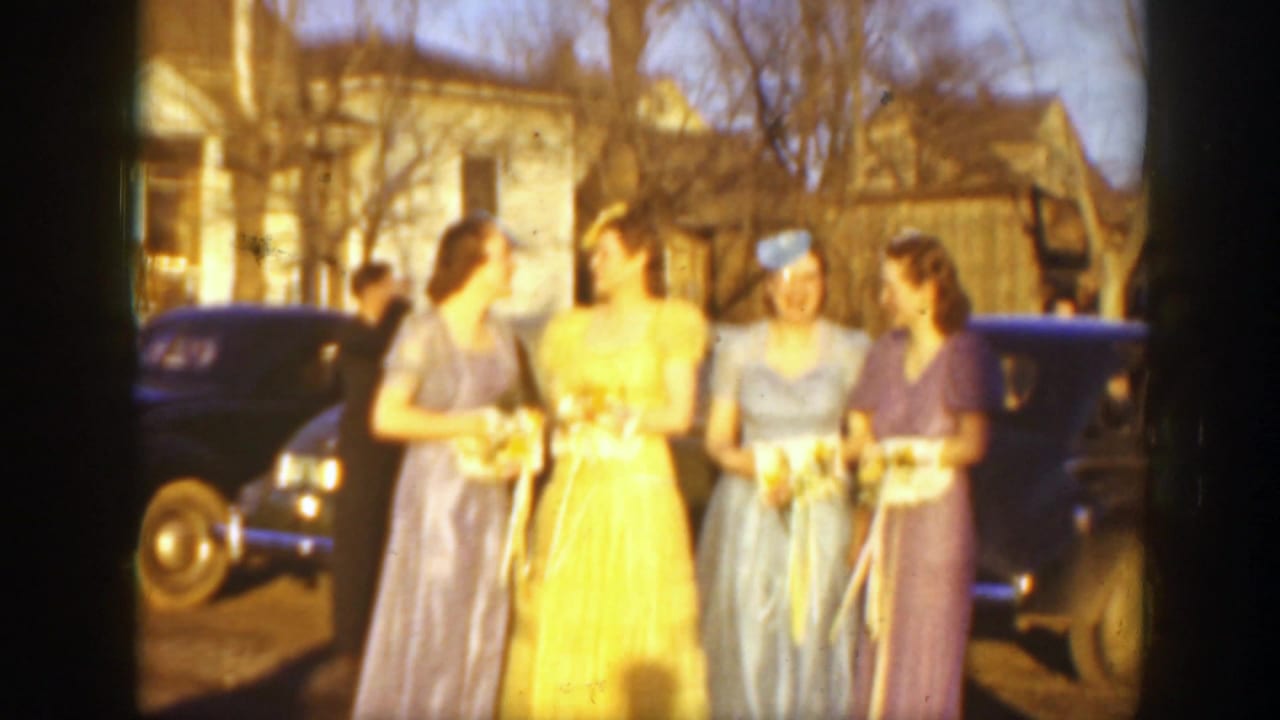 1939 Montana home-movie frame of four young women, likely bridesmaids, posing with bouquets in front of dark automobiles and houses.