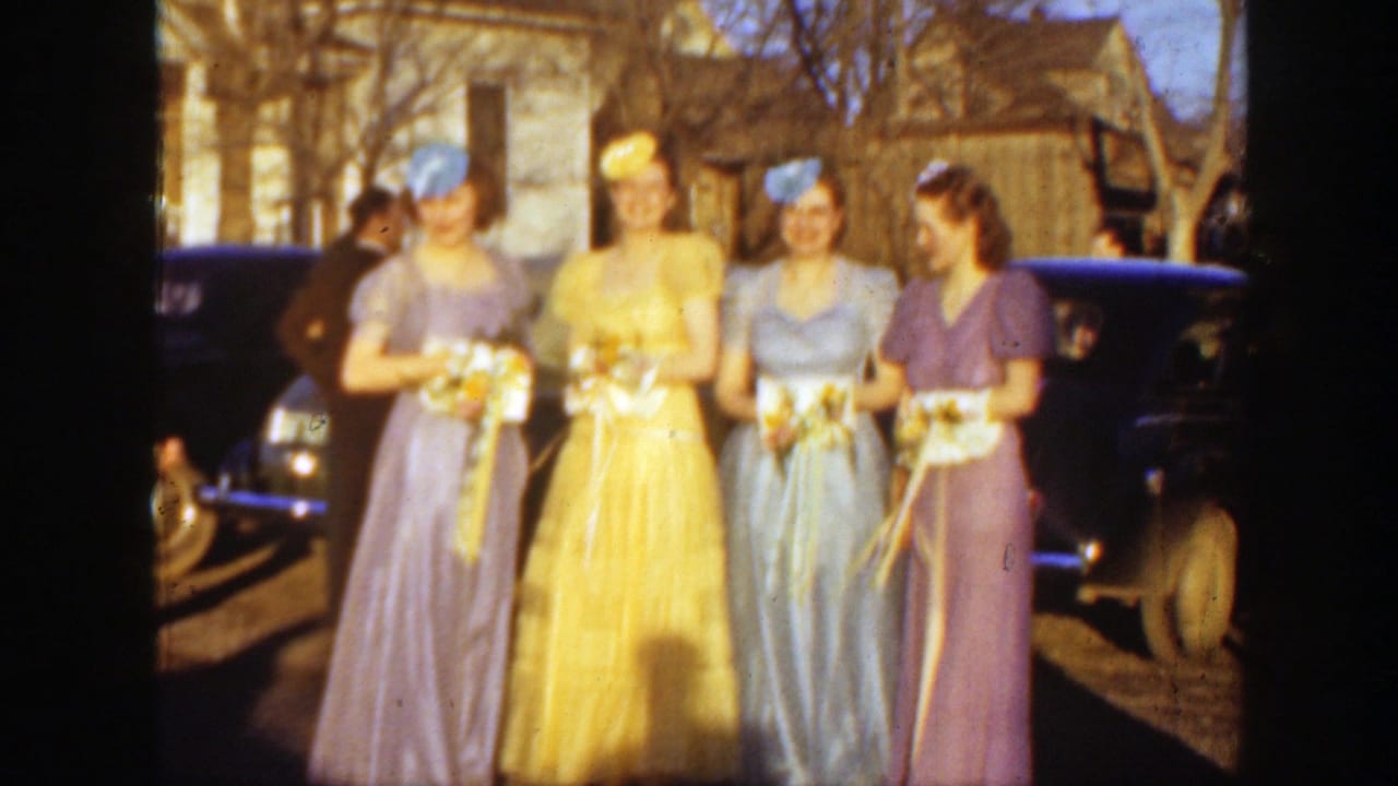 1939 Montana frame showing four young women, likely bridesmaids, posing with bouquets before a dark car and residential buildings.