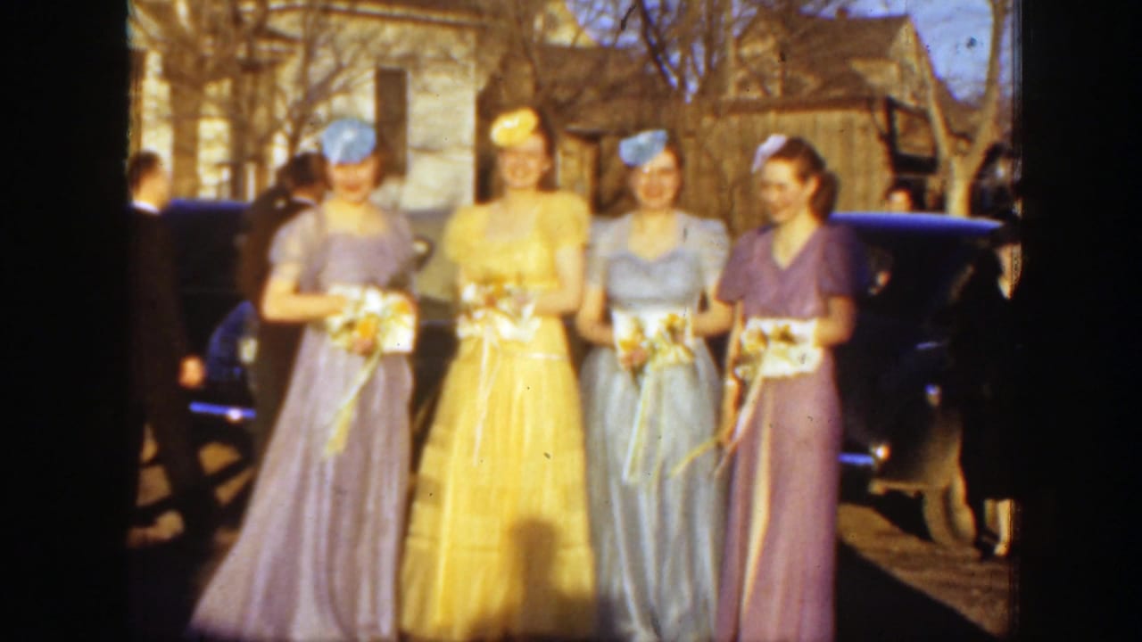 Four women in pastel gowns hold bouquets outdoors near parked cars.