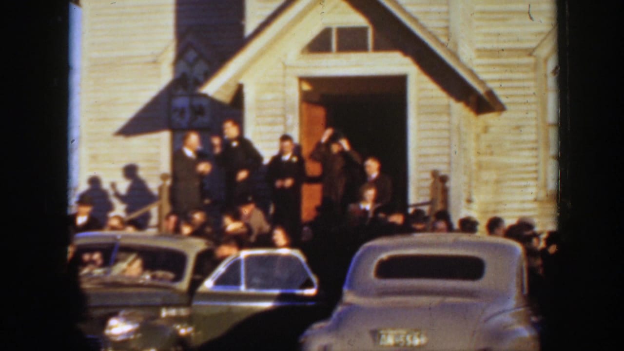 A crowd stands on the steps of a white clapboard church in Montana, with late-1930s cars parked along the street.