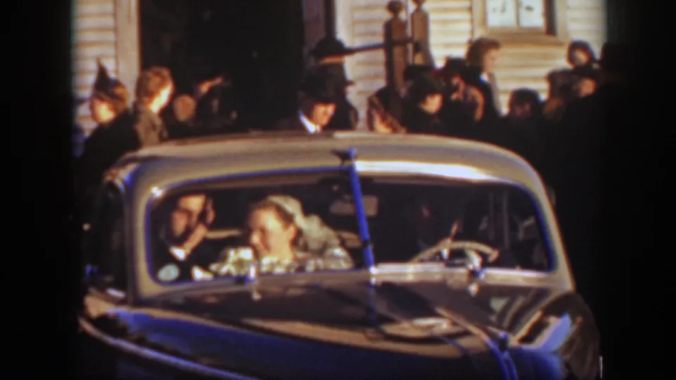 The bride and groom smile inside a ribboned car as guests crowd around the church exterior in 1939 Montana footage.