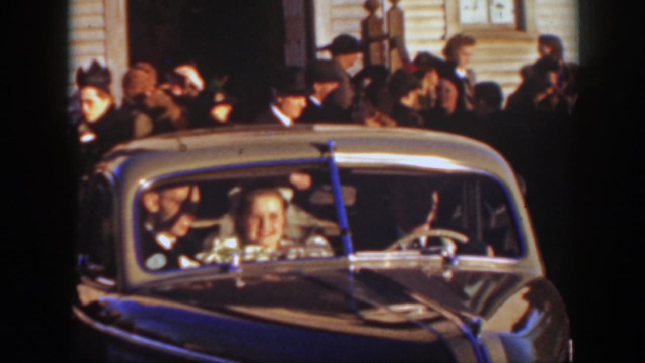 Bride and groom seated in a classic car with guests crowded behind them.