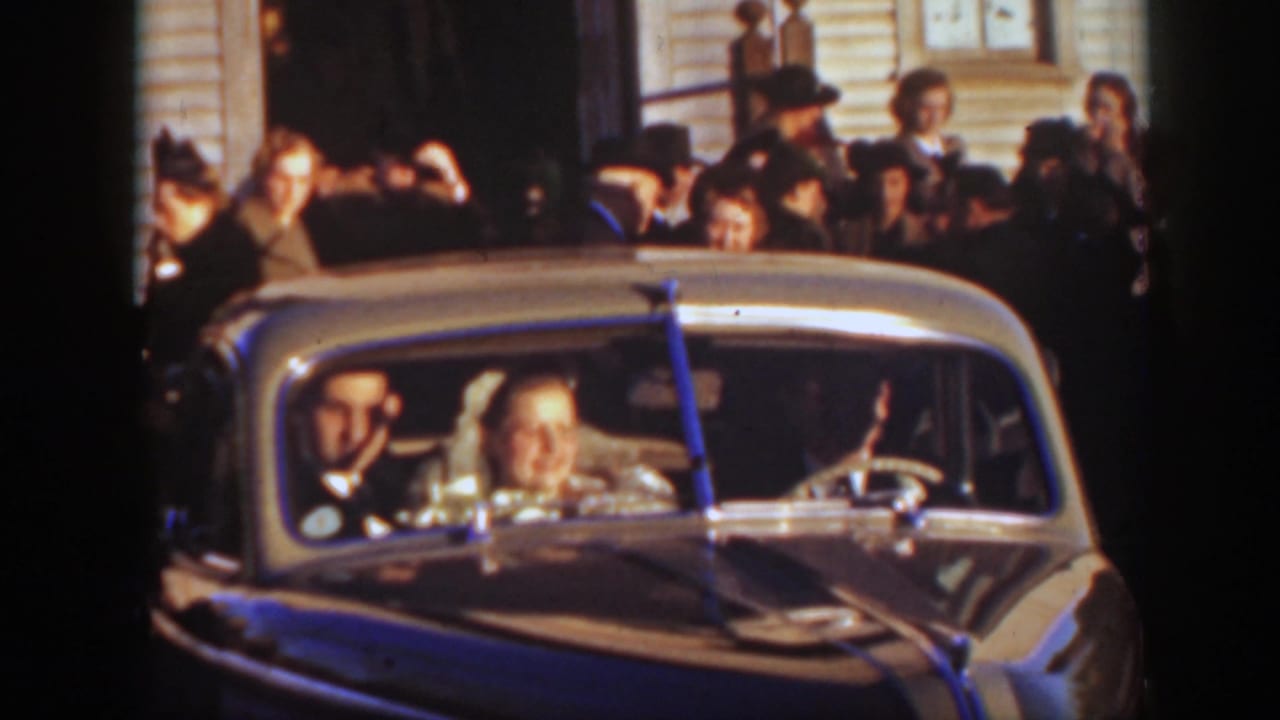 The couple smiles from inside a rounded classic car as a crowd in formal 1930s clothing stands behind them in Montana.