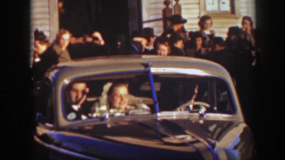 The couple smiles from inside a rounded classic car as a crowd in formal 1930s clothing stands behind them in Montana.