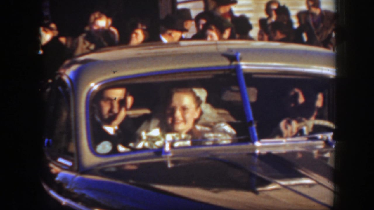 Bride and male companion seated in a light-colored vintage car with onlookers behind them.