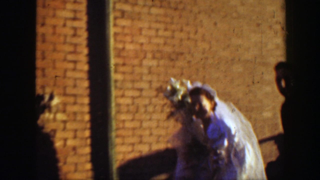 Bride in a white gown and veil walks beside a dark brick wall in dim light.