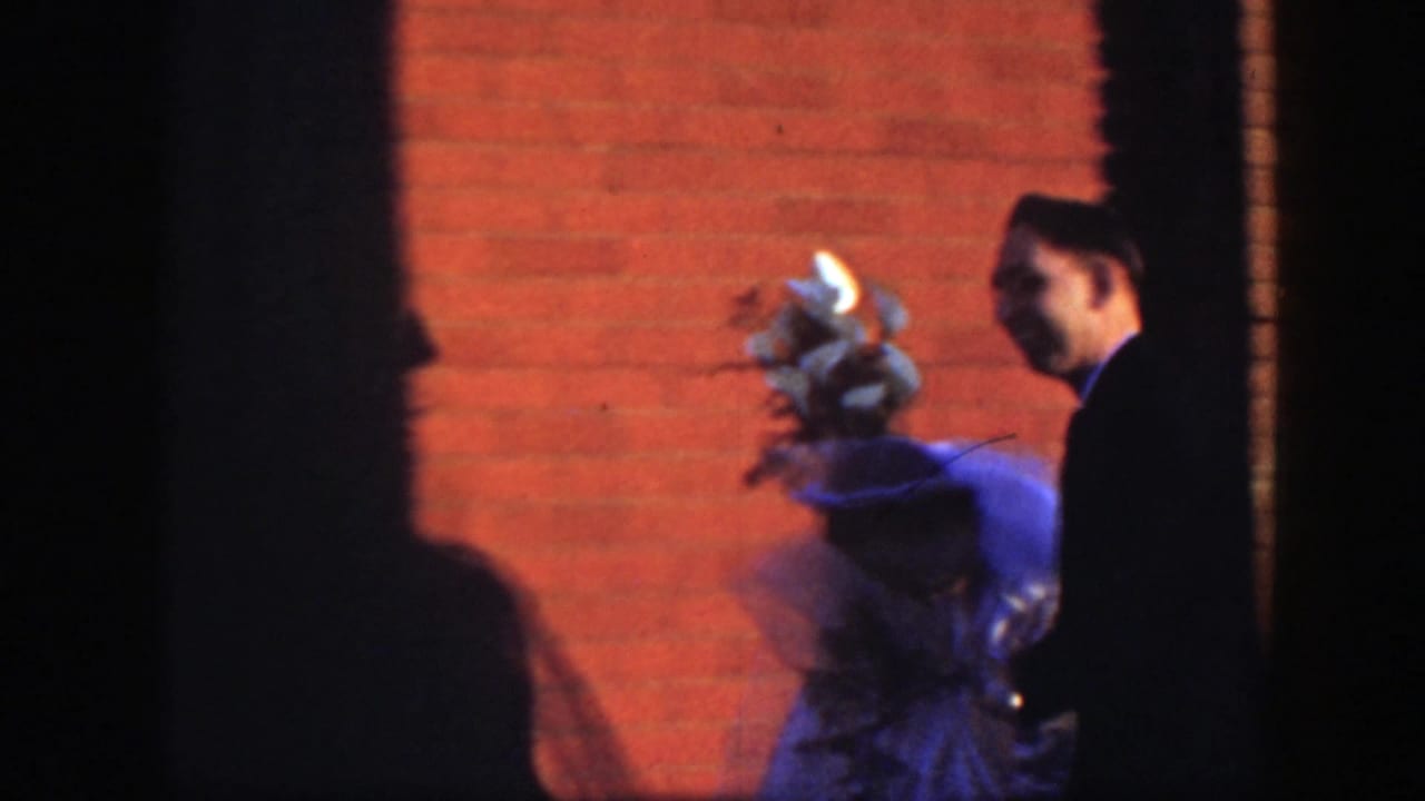 A groom in a dark suit appears near the bride and bouquet, with a red brick wall and heavy shadow in the background.
