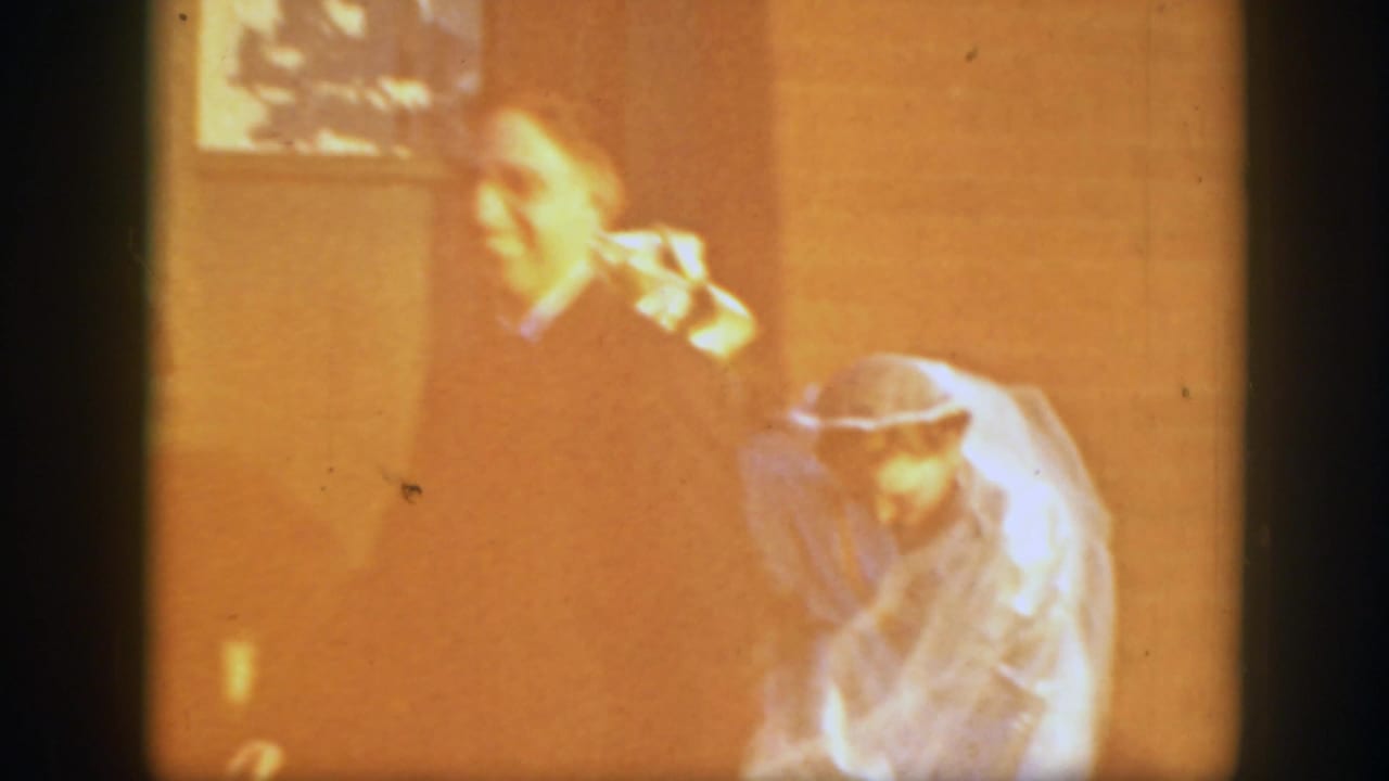 The bride appears to bend or adjust her gown while another adult figure stands nearby in soft, yellowish 8mm light.