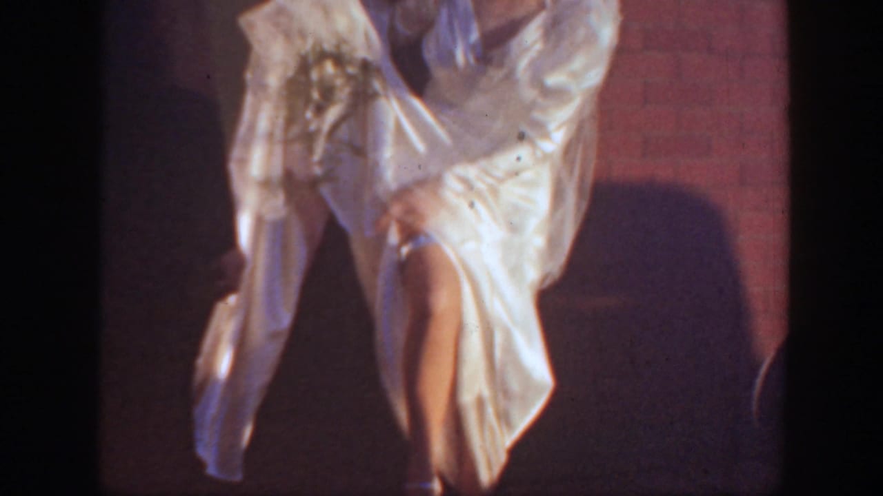 A close, shadowed view shows the bride's white gown and leg as she appears to lift or adjust the dress against a brick wall.