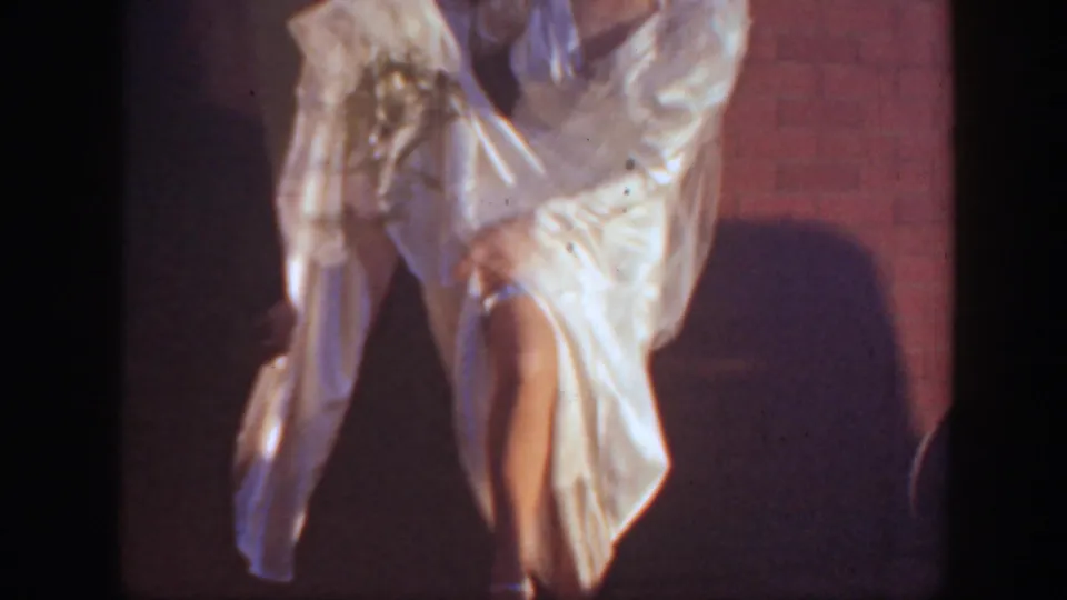 A close, shadowed view shows the bride's white gown and leg as she appears to lift or adjust the dress against a brick wall.