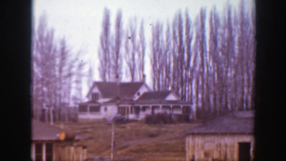 1939 Montana home-movie frame showing a house, open yard, small outbuilding, and tall leafless trees in soft focus.