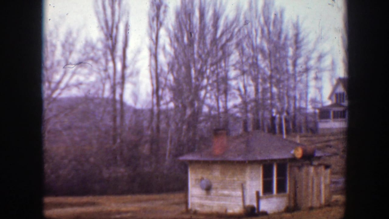 1939 Montana home-movie frame showing a small cottage or outbuilding beneath bare trees on a quiet hillside.