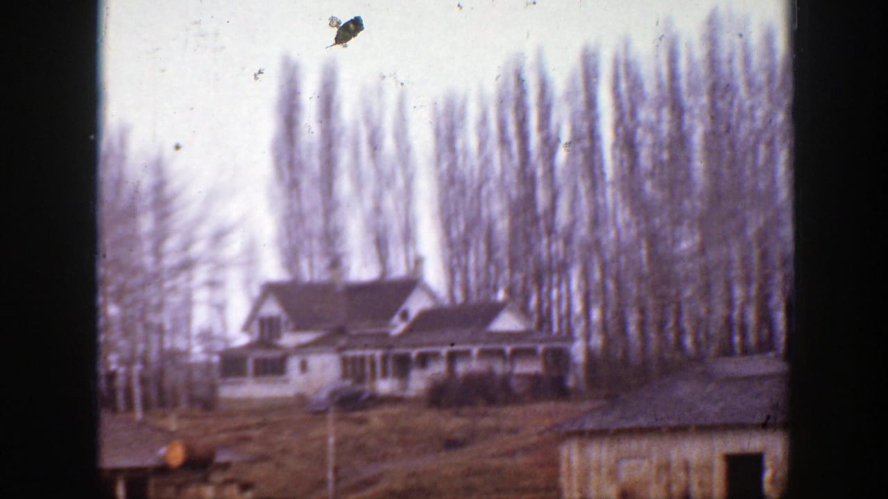 1939 Montana frame showing a hillside house, adjacent outbuilding, bare trees, and open ground under an overcast sky.