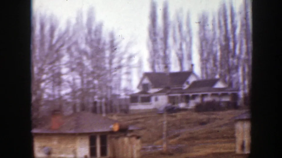 1939 Montana 8mm frame showing a farmhouse on a rise, a small outbuilding in front, and tall bare trees in the background.