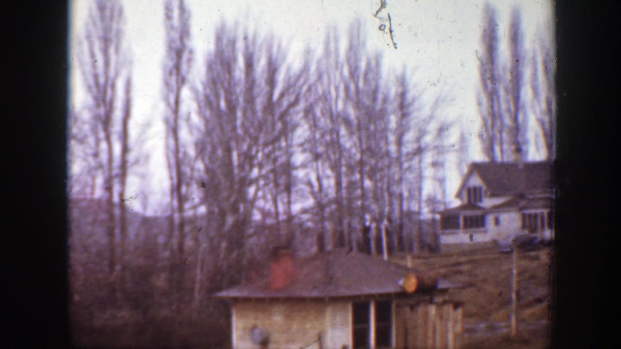 1939 Montana farm view with a house, small outbuilding, bare tree line, and open ground in a soft home-movie frame.