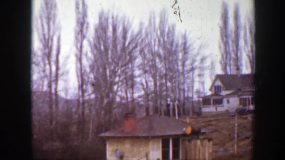 1939 Montana farm view with a house, small outbuilding, bare tree line, and open ground in a soft home-movie frame.