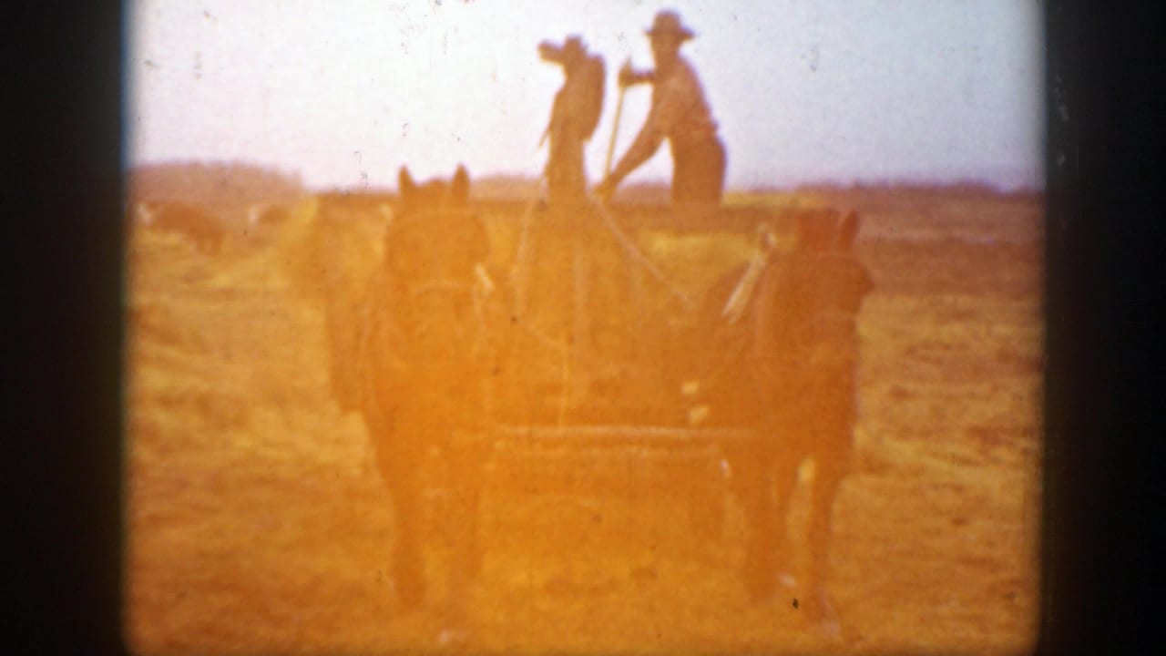In 1939 Montana home-movie footage, two farmers in hats work atop a hay wagon in a dry open field.