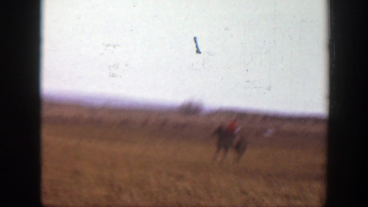 Small rider on horseback crossing a dry open field under a pale sky.